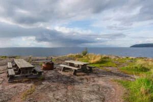 Grillplats med två träbänkar och eldstad på klipphällar vid Vättern i Stora Lunds naturreservat, under dramatisk molnhimmel