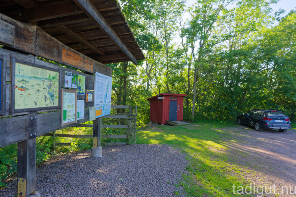 Informationstavla med kartor och skyltar vid parkeringen i Kråkeryd naturreservat, med röd toalettbyggnad och lövskog i bakgrunden.