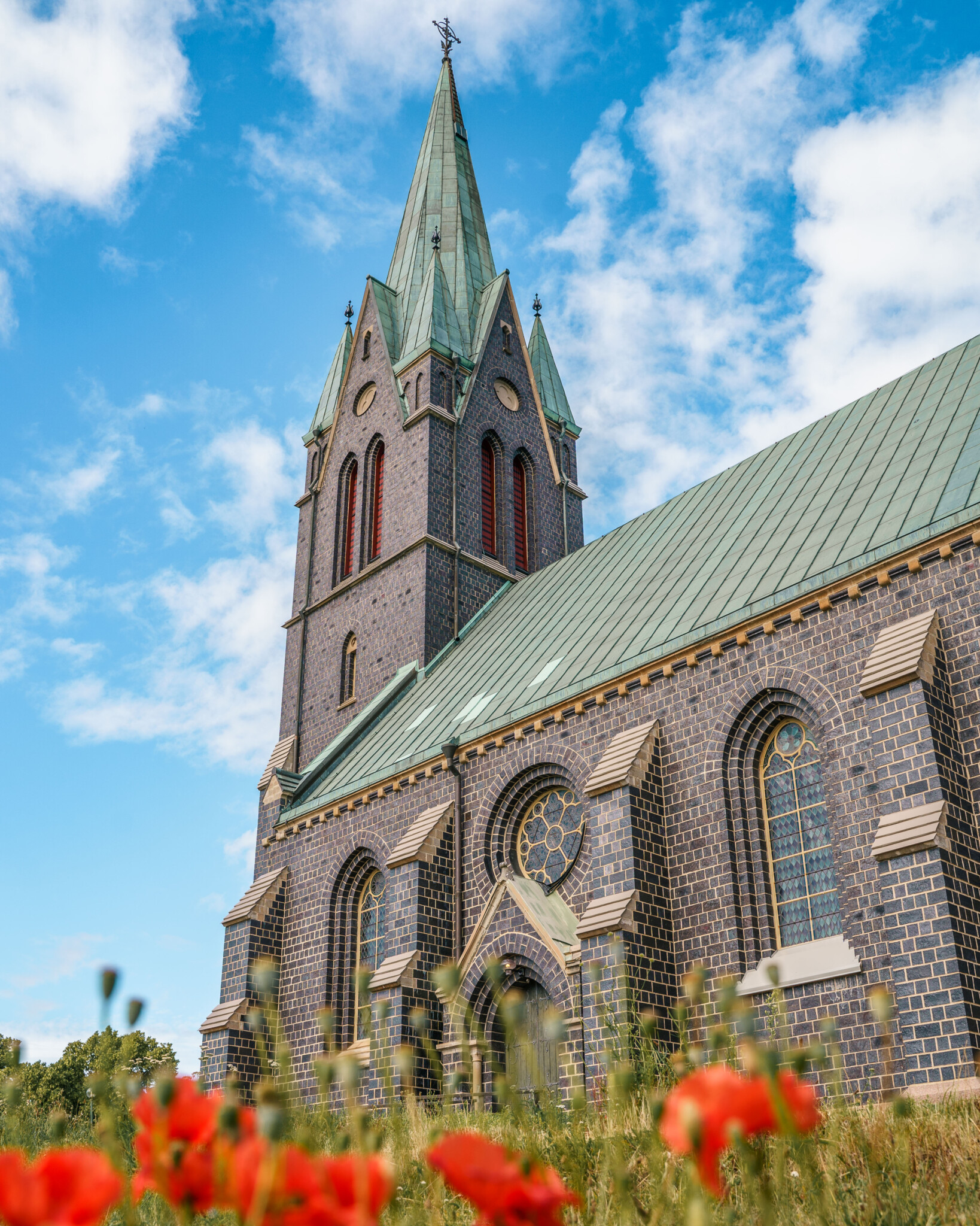 Nygotisk kyrka i mörkt tegel med grönpatinerat koppartak och högt torn, omgiven av röda vallmor en sommardag i Boxholm.