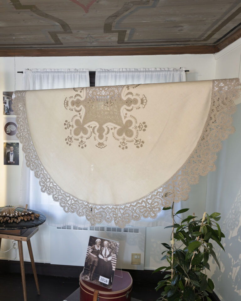 Stor handgjord spetsduk med dekorativt mönster upphängd på vägg i Vadstena Spetsmuseum, med knypplingsverktyg och fotografier i rummet.