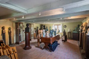 Utställningsrum på Skulpturmuseet Gottfrid Larsson-gården i Vadstena med skulpturer och byster på piedestaler och ett stort bord, tegelgolv och vitkalkat bjälktak.