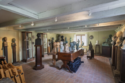 Utställningsrum på Skulpturmuseet Gottfrid Larsson-gården i Vadstena med skulpturer och byster på piedestaler och ett stort bord, tegelgolv och vitkalkat bjälktak.