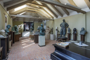 Utställningsrum på Skulpturmuseet Gottfrid Larsson-gården i Vadstena med bronsskulpturer, byster och figurer under synliga takbjälkar