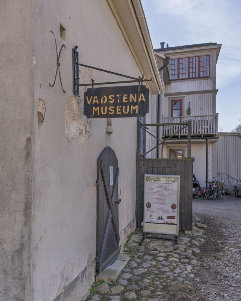 Entrén till Vadstena Museum med en smidesskylt i guldbokstäver, kullersten och en öppen träport mot en innergård.