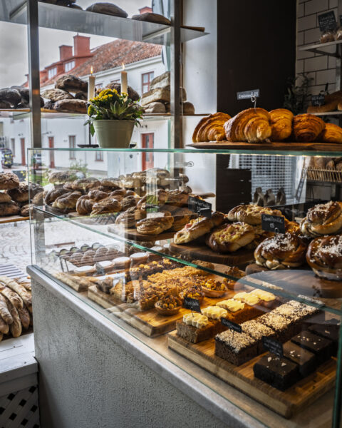 Glasdisk fylld med nybakade kanelbullar, croissanter, kakor och bröd på Bageri Hamilton i Vadstena, med vita stadshus synliga genom fönstret