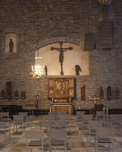 Interiör i Vadstena Klosterkyrka med medeltida krucifix, förgyllt altarskåp, stenväggar, kristallkrona och vita stolar