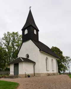 Nässja kyrka med vitputsade väggar, mörkt spåntak och tornspira med kors, omgiven av lövträd på Östgötaslätten.
