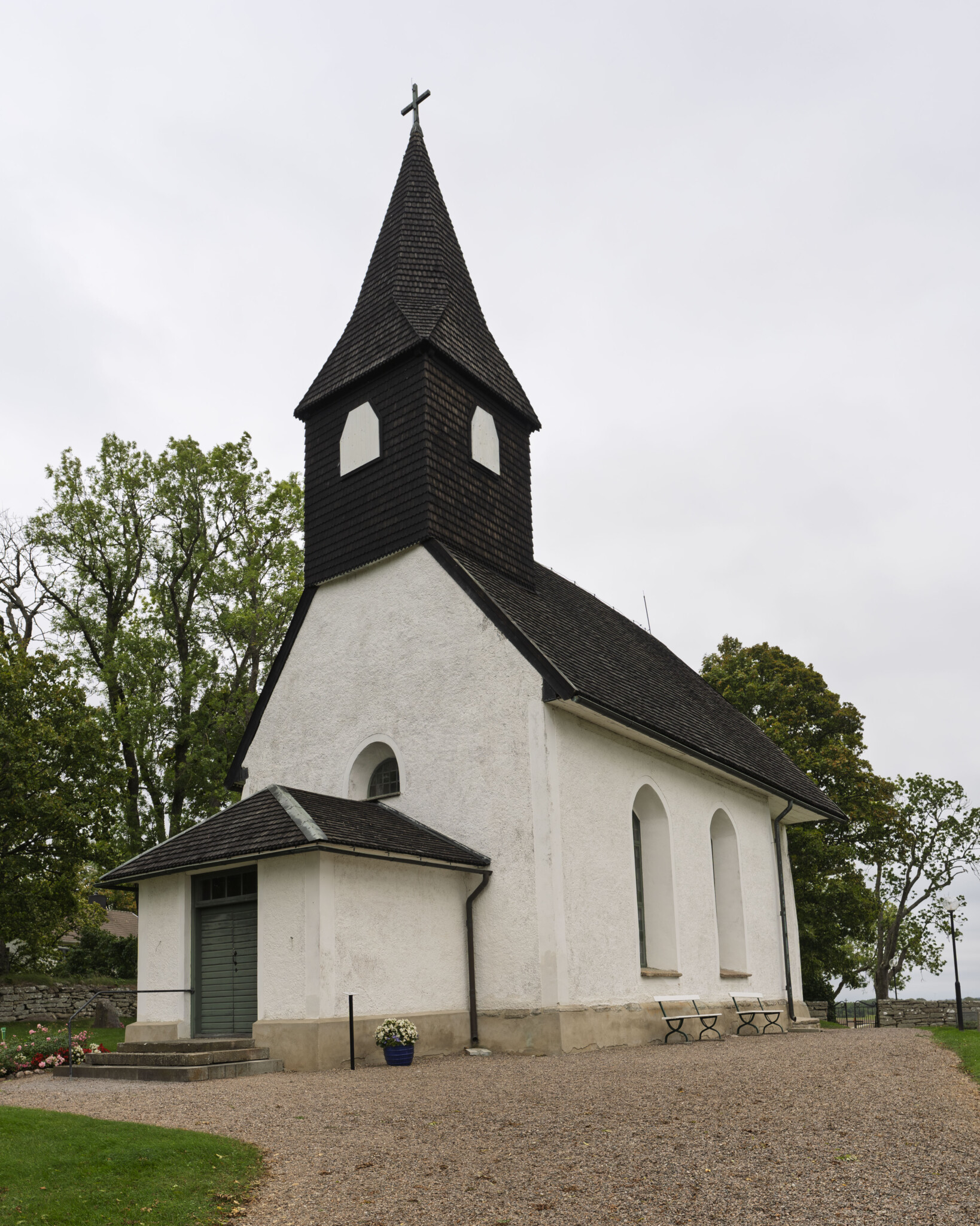 Nässja kyrka med vitputsade väggar, mörkt spåntak och tornspira med kors, omgiven av lövträd på Östgötaslätten.