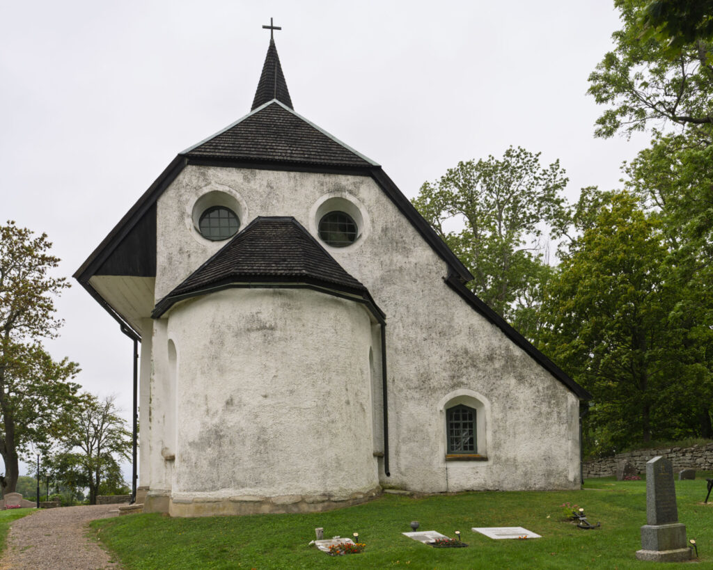 Nässja kyrka, en vitputsad medeltidskyrka med mörkt tak och kors på tornspiran, omgiven av kyrkogård och gröna lövträd.