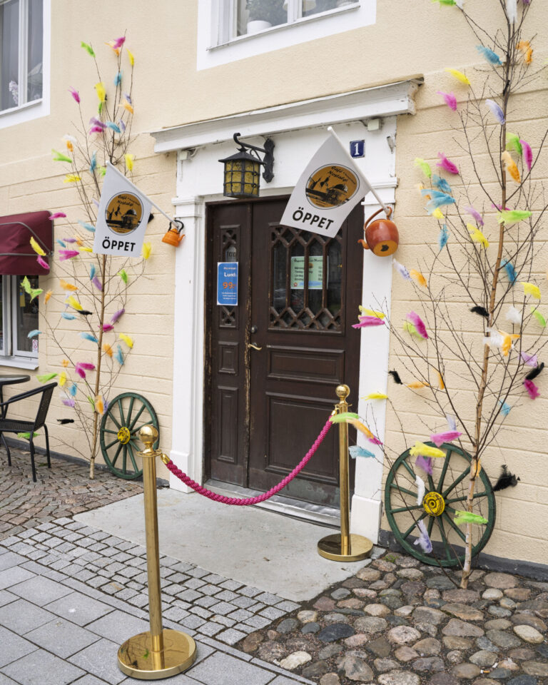 Entré till Café Nostalgi i Vadstena med brun trädörr, "Öppet"-skyltar, avspärrningsrep i mässing och påskris med färgglada fjädrar längs fasaden.