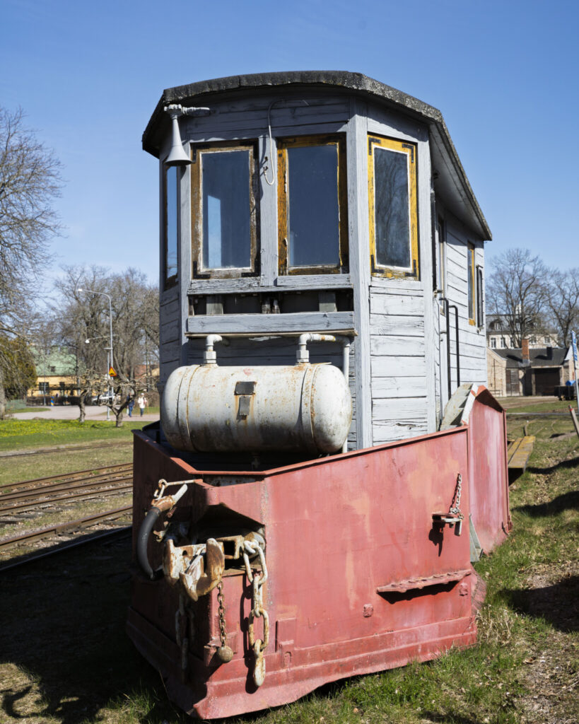 Gammalt smalspårigt lok med röd front och grå trähytt vid Wadstena-Fogelsta Järnväg i Vadstena, en solig vårdag