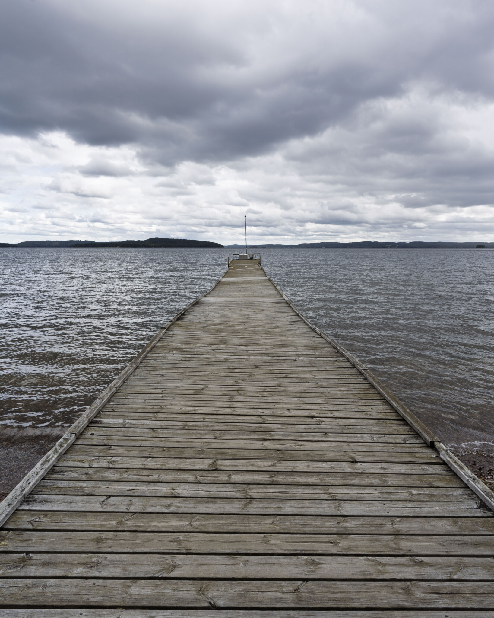 Lång träbrygga som sträcker sig rakt ut i sjön Sommen vid Malexander en mulen dag, med dramatisk molnhimmel och kullar i horisonten.