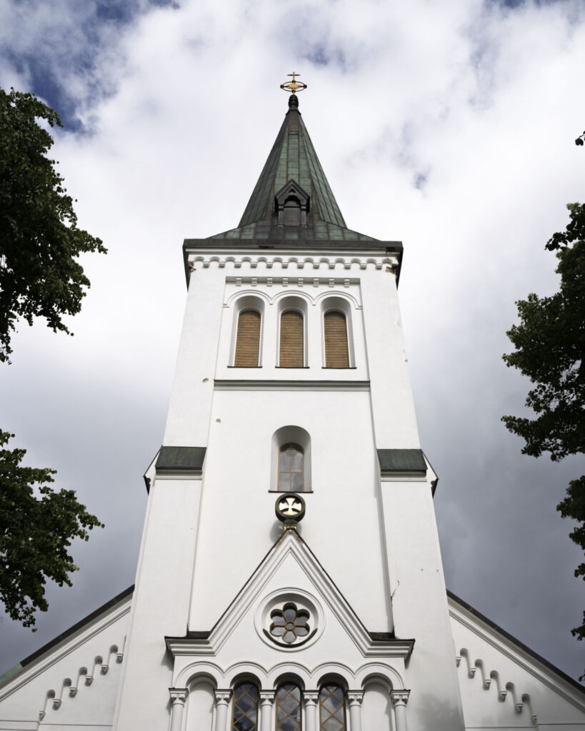 Malexander kyrkas vitputsade torn med spetsig grön spira och gyllene kors, omgivet av lövträd mot en molnig himmel.