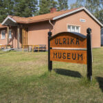 Träskylt med texten "Ulrika Museum" framför en rosa putsad byggnad med tegeltak, omgiven av gräsmatta och träd en solig dag.