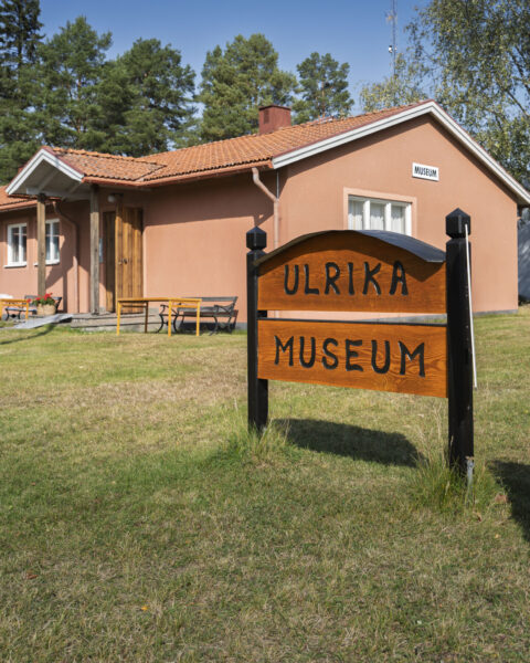 Träskylt med texten "Ulrika Museum" framför en rosa putsad byggnad med tegeltak, omgiven av gräsmatta och träd en solig dag.