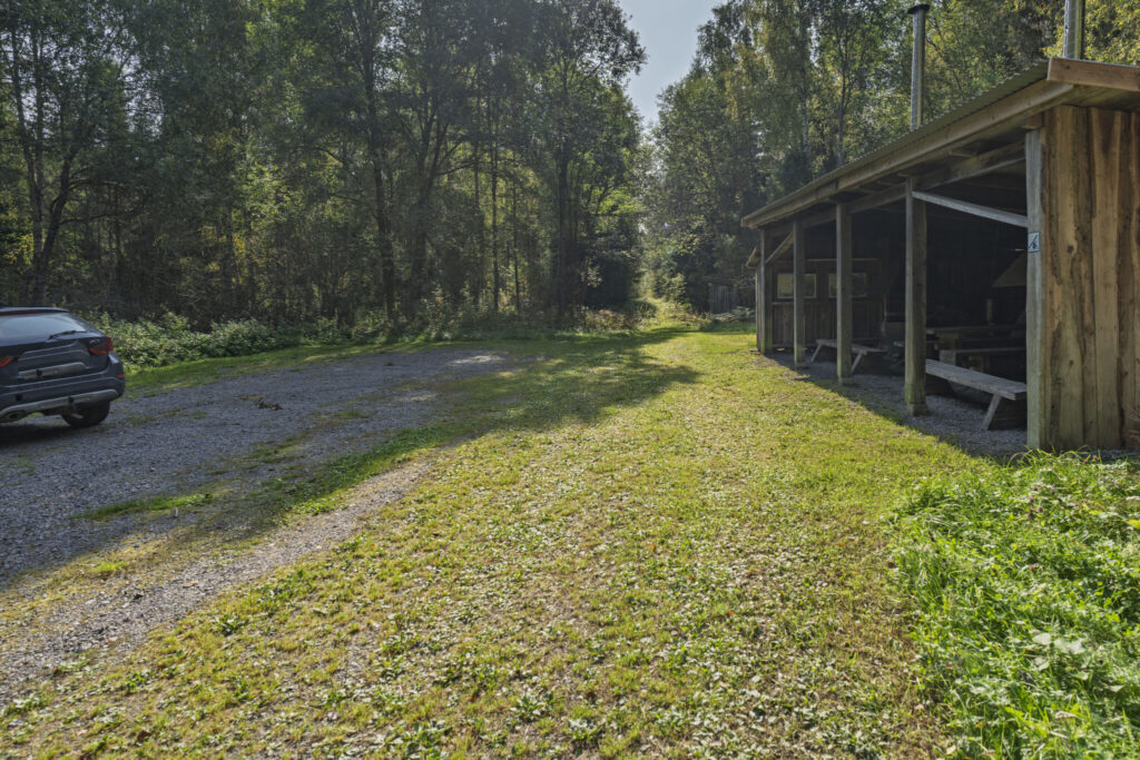 Parkering vid Ycke urskog med grusplan, parkerad bil och öppen rastplats under tak med bänkbord, omgiven av lummig lövskog.