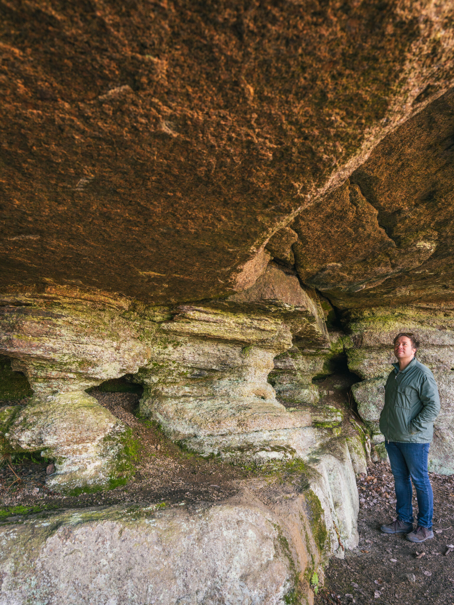 Man står under ett massivt klippöverhäng i Pelargrottan vid Pelarberget i Malexander, med vittrade sandstensformationer och mossbeklädda pelare.