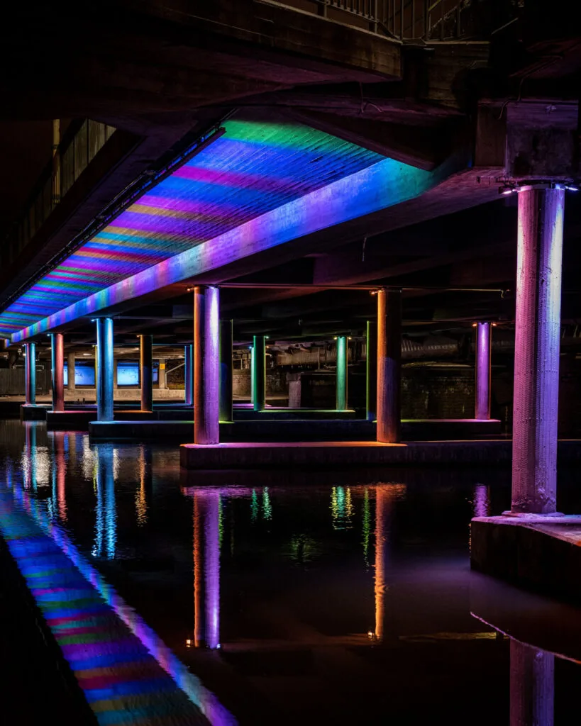 Färgglad ljusinstallation under en bro med pelare i regnbågsfärger som speglas i det stilla vattnet nedanför