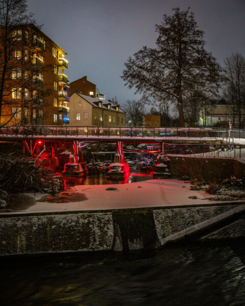 Vinterkväll vid en å med röd ljusbelysning under en gångbro, snötäckta stenar och flervåningshus med upplysta fönster i bakgrunden.