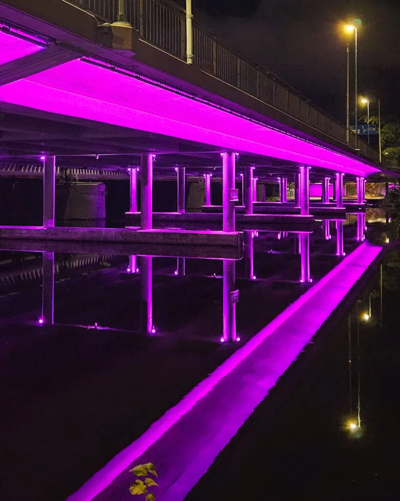 Bro upplyst med lila LED-belysning nattetid, med pelare som speglas i det stilla vattnet undertill