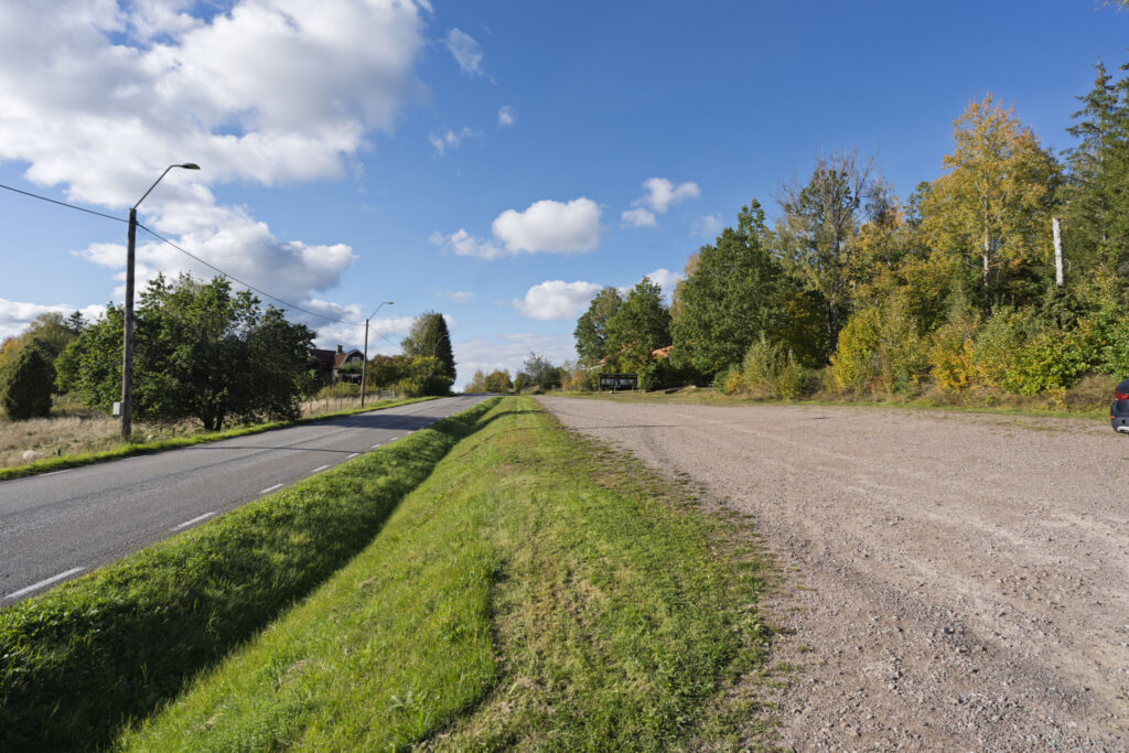 Landsväg med grönt dike vid Kulladal nära Västra Hargs naturreservat, med lövträd i höstfärger och blå himmel med vita moln.