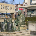 Bronsskulptur av en sagoberättare som läser för tre barn i Mjölby centrum, med stadsmiljö i bakgrunden.