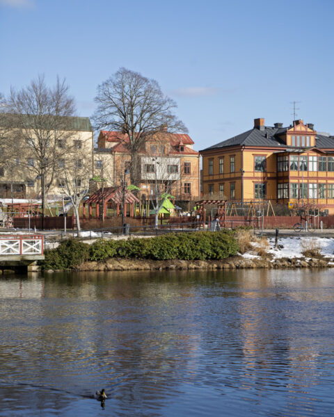 Stadsvy över centrala Mjölby med färgglada byggnader, lekplats och park längs Svartån en tidig vårdag med snörester och en and i vattnet
