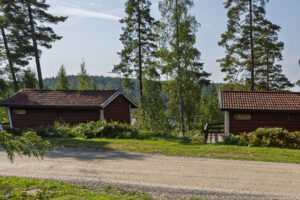 Röda stugor med skyltar vid en grusväg omgivna av tallar och grönska nära en sjö längs Östgötaleden i Östergötland