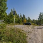 Parkeringsplats vid Amundeboslingan och Drögenslingan på Östgötaleden med vägvisningsskyltar, en parkerad bil och grusväg omgiven av björk- och barrskog.
