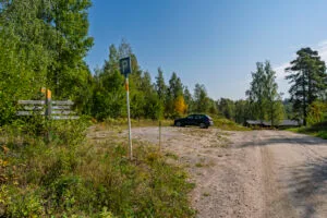 Parkeringsplats vid Amundeboslingan och Drögenslingan på Östgötaleden med vägvisningsskyltar, en parkerad bil och grusväg omgiven av björk- och barrskog.