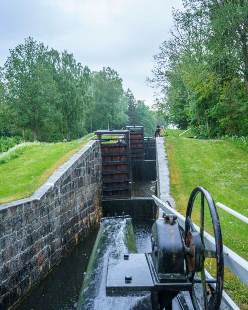 Slussport med vevmekanism vid Hovetorps slussar längs Kinda kanal, omgiven av gröna gräsvallar och lövträd en mulen dag.