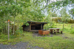 Vindskydd i trä med grillplats och sittbänk vid Hovetorps slussar längs Östgötaleden, omgivet av lummig grönska