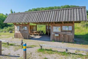 Rastplats i trä med tak, sittbänk och informationstavlor vid Viggebysandsbadet, omgiven av grönska och med ledmarkering i förgrunden.