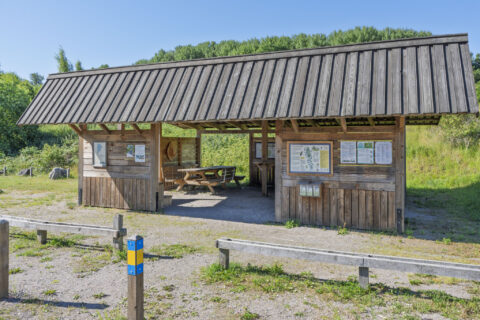 Rastplats i trä med tak, sittbänk och informationstavlor vid Viggebysandsbadet, omgiven av grönska och med ledmarkering i förgrunden.