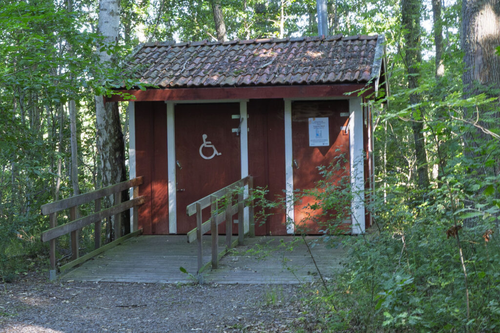 Röd handikappanpassad utedass med ramp vid Viggebysandsbadet, omgivet av lövskog i Viggeby naturreservat