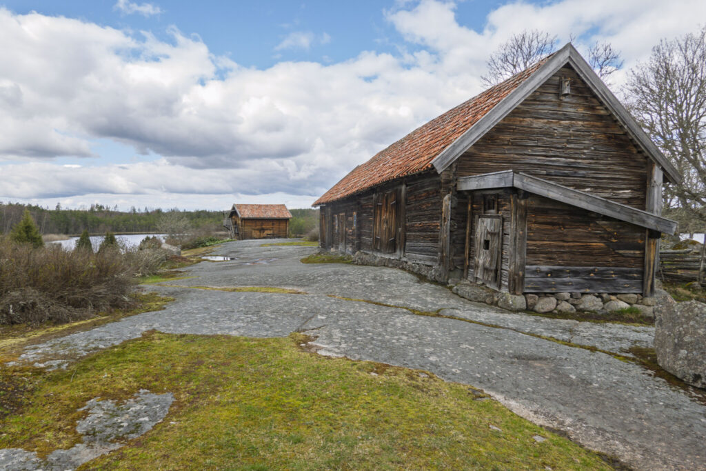Gamla timmerbyggnader med tegeltak vid kulturreservatet Öna, omgivna av berghällar, mossa och norrländskt landskap med sjö i bakgrunden
