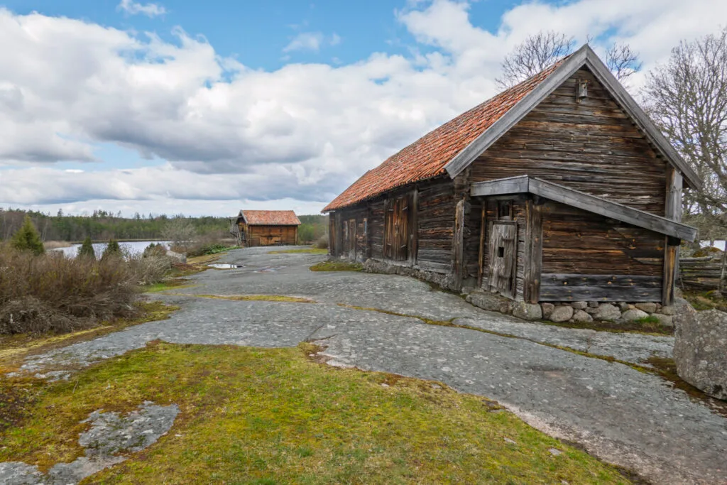 Gamla timmerbyggnader med tegeltak vid kulturreservatet Öna, omgivna av berghällar, mossa och norrländskt landskap med sjö i bakgrunden