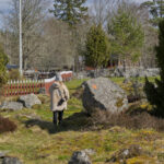 Person promenerar bland stora stenblock och grönska vid Öna kulturreservat i Östergötland, med rött staket och flaggstång i bakgrunden.