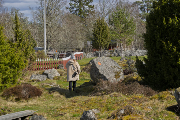 Person promenerar bland stora stenblock och grönska vid Öna kulturreservat i Östergötland, med rött staket och flaggstång i bakgrunden.