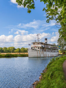 Kanalbåten M/S Wilhelm Tham på Göta kanal en solig sommardag, med svensk flagga i aktern och gröna träd längs dragvägen.