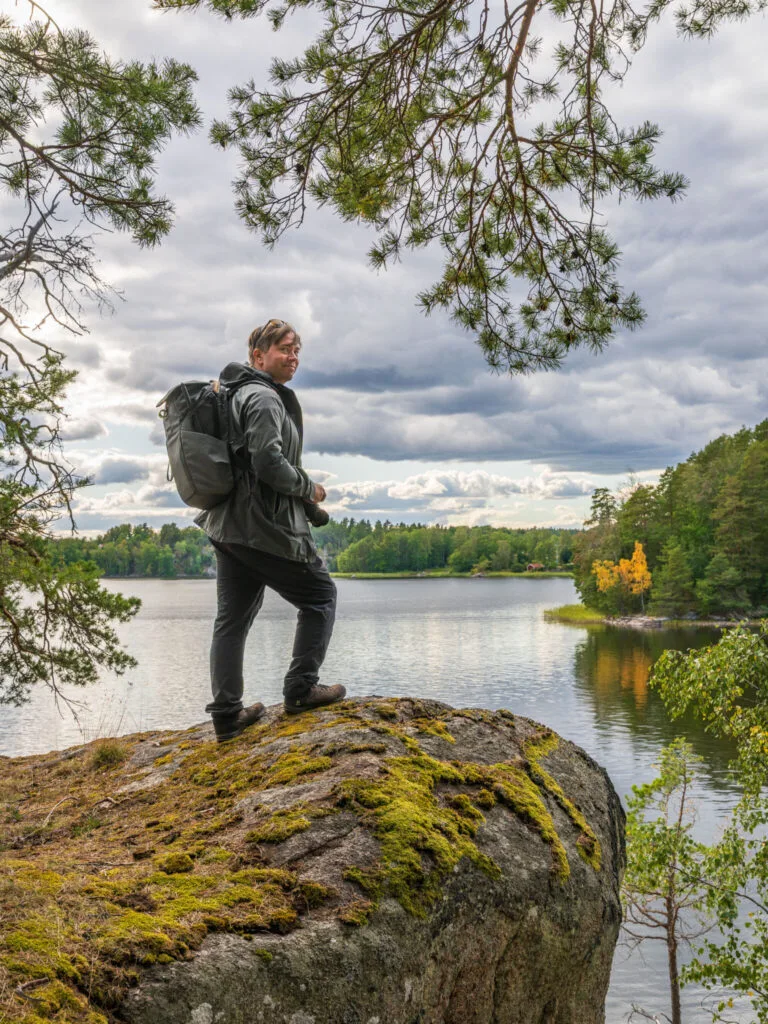 Vandrare med ryggsäck står på mossbetäckt klippa vid lugnt vatten längs Viggebyleden i Linköping.