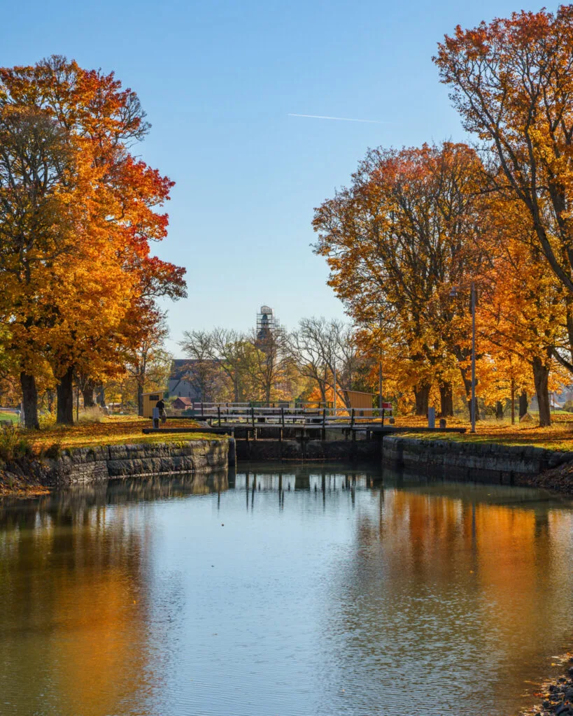 Göta kanal en höstdag med färgsprakande träd i orange och rött, en sluss och stilla vattenspeglar under klarblå himmel.