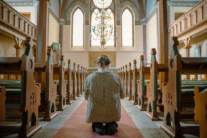 Kvinna knäböjer i mittgången i Orlunda kyrka med glasmålningar, kristallkrona och vackert dekorerat kor i bakgrunden