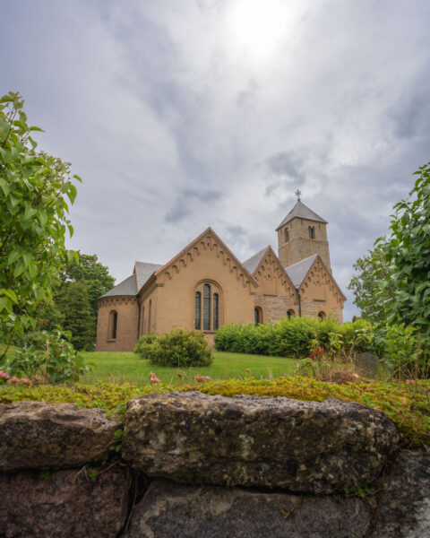 Heda kyrka i Ödeshög sedd från kyrkogårdsmuren, med romanskt stentorn och gulputsade fasader omgivna av grönska.