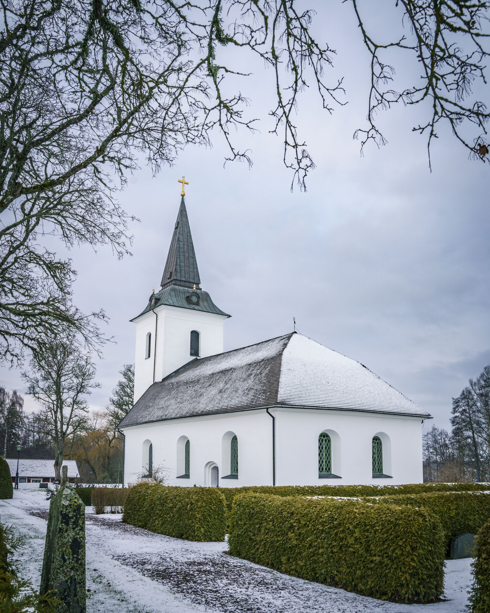 Sya kyrka i Mjölby kommun en vinterdag med lätt snö på marken, omgiven av kyrkogård med klippta häckar och kala träd.