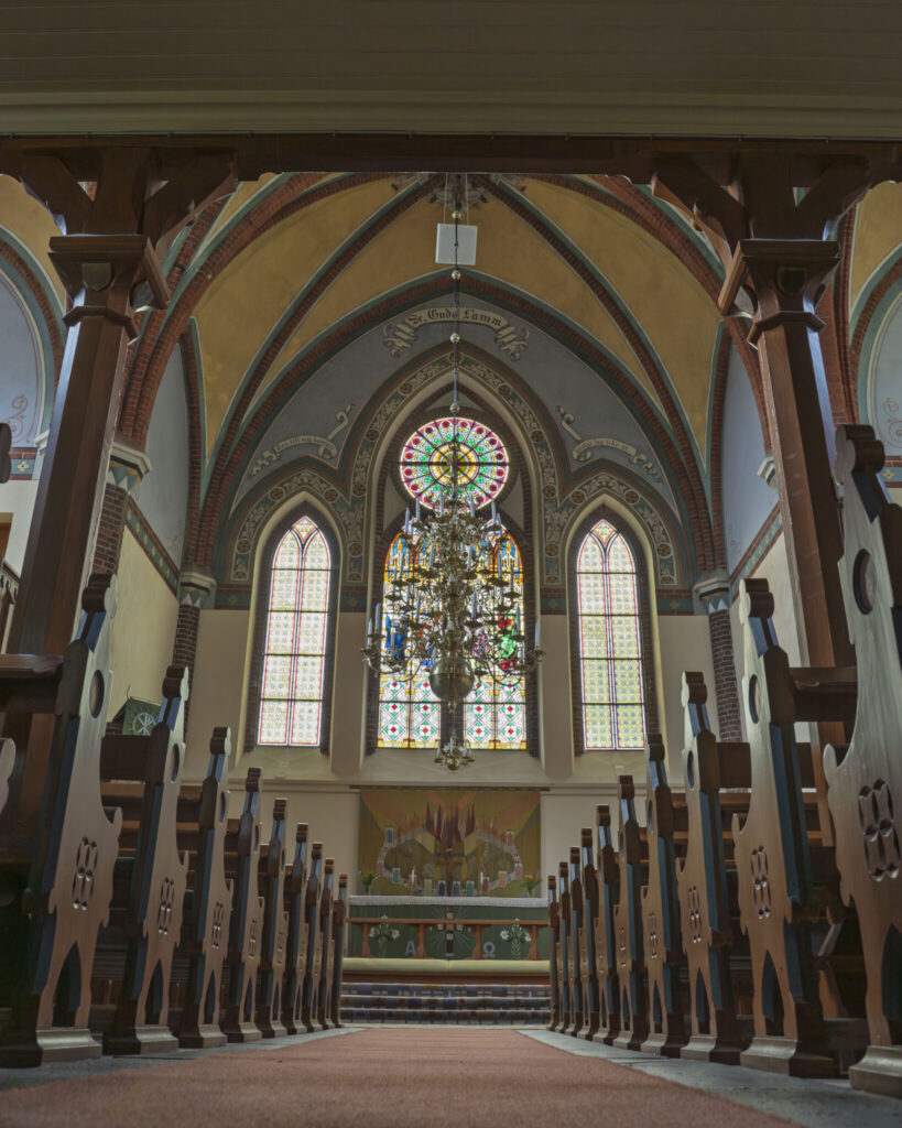 Interiör i Orlunda kyrka med nygotiska valv, färgglada glasmålningar, stor ljuskrona och träbänkar längs mittgången mot altaret.