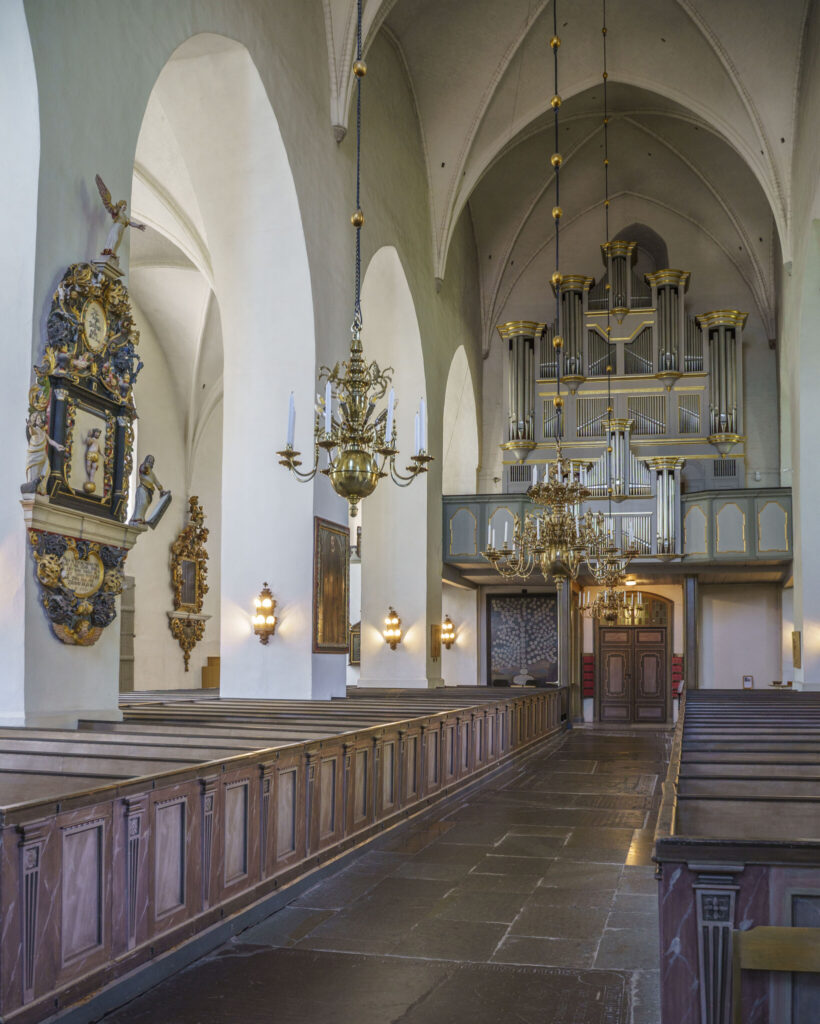 Interiör i Vårfrukyrkan i Skänninge med vita gotiska valv, träbänkar, mässingskronor och orgel vid västra väggen.
