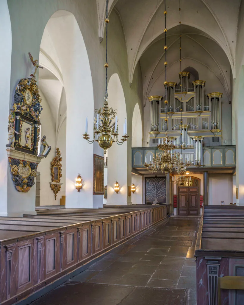 Interiör i Vårfrukyrkan i Skänninge med vita gotiska valv, träbänkar, mässingskronor och orgel vid västra väggen.