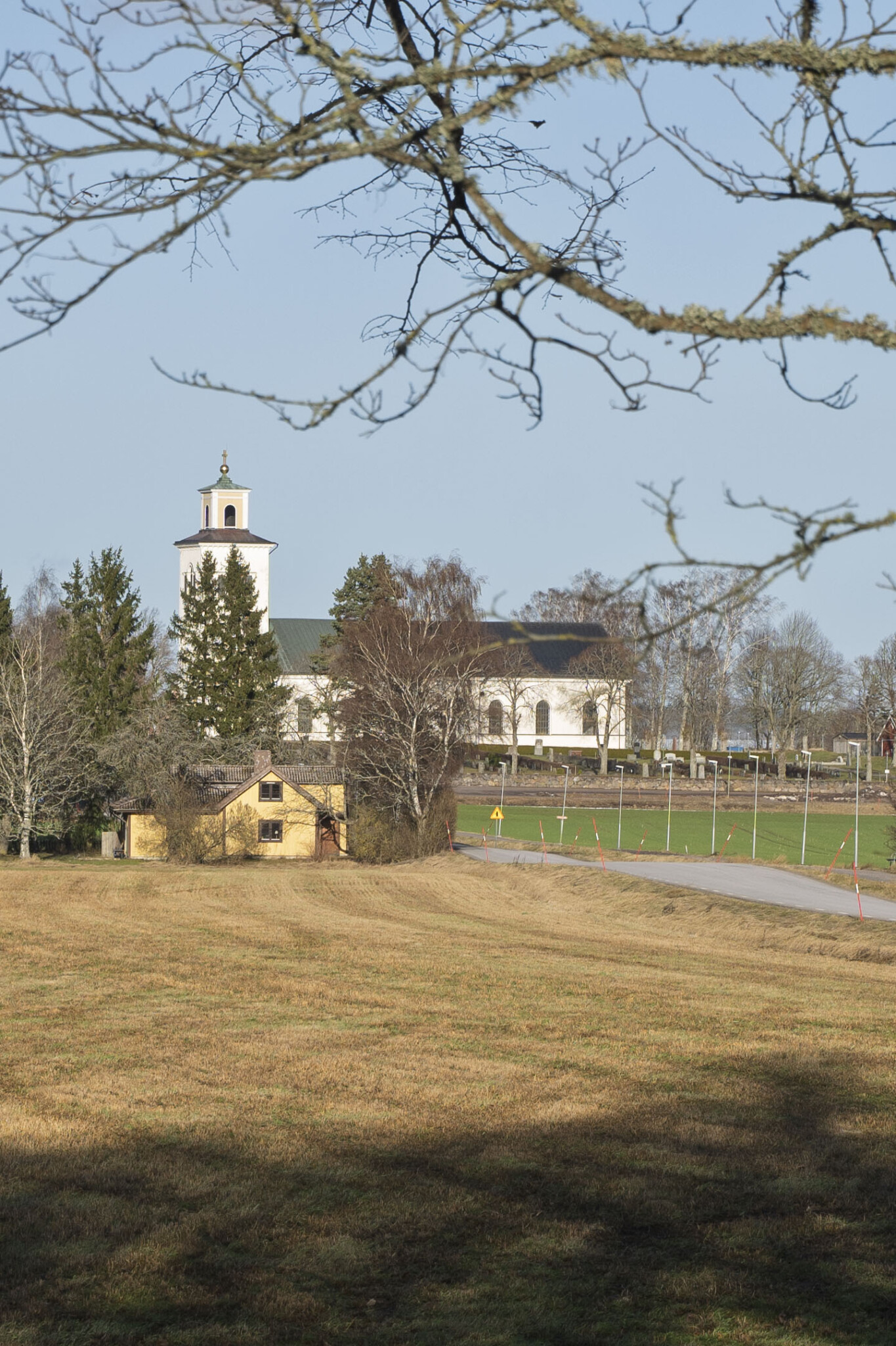 Klockrike kyrka i Motala kommun, en vitputsad kyrka med torn omgiven av öppna fält, en gul stuga och kala träd i tidig vår.