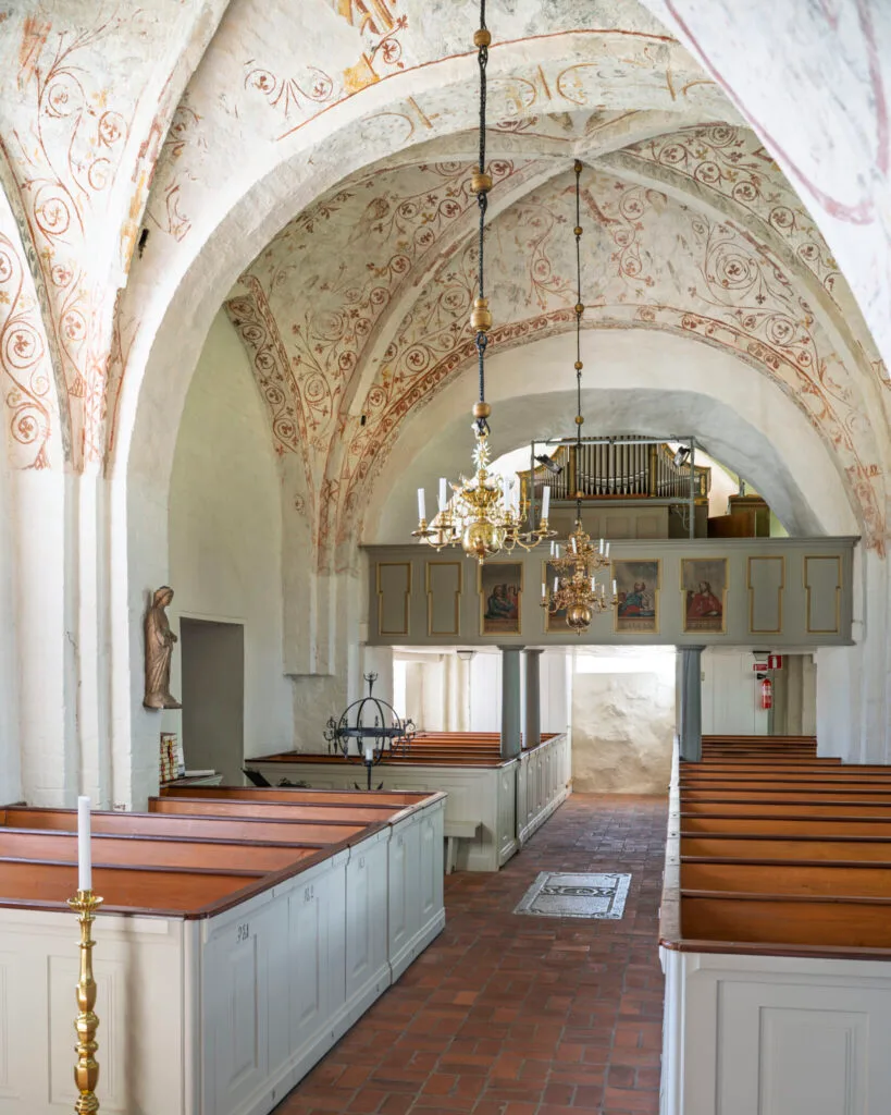Interiör i Järstads kyrka med medeltida kalkmålningar på kryssvalven, kyrkbänkar i trä, mässingskronor och orgelläktare i bakgrunden